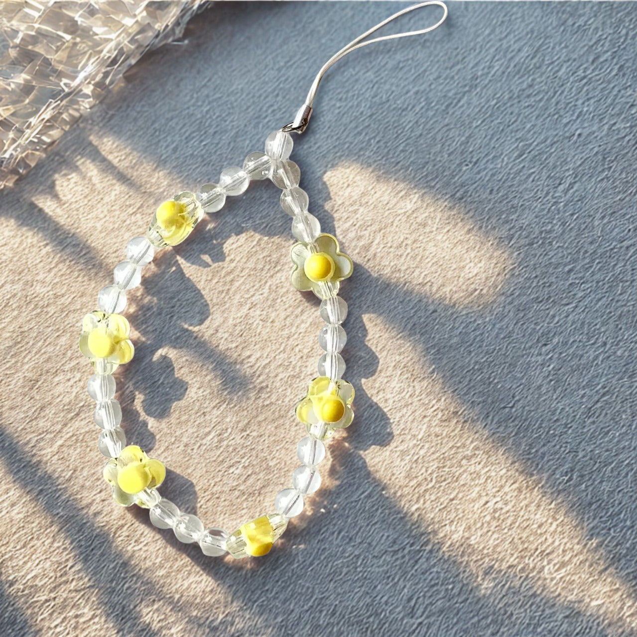 A yellow and clear beaded floral phone charm placed on a grey surface, illuminated by warm, dappled sunlight and shadows.