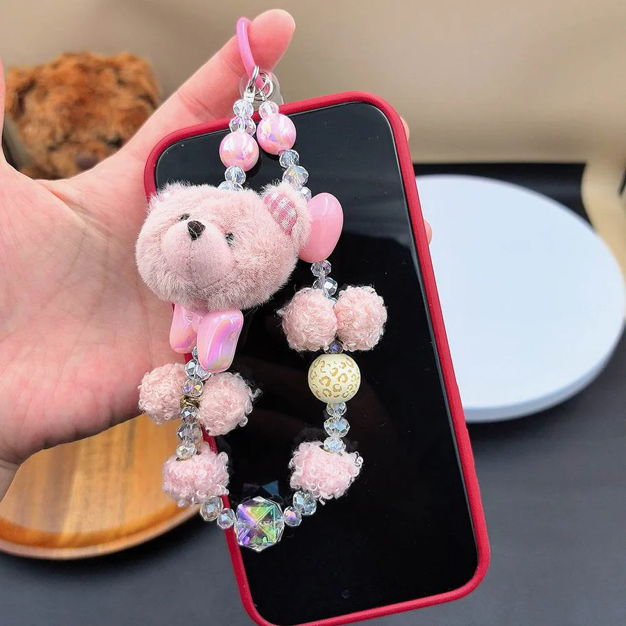Pink plush bear phone charm featuring fluffy pom-pom beads, clear crystals, and an iridescent bow attached to a black smartphone case.
