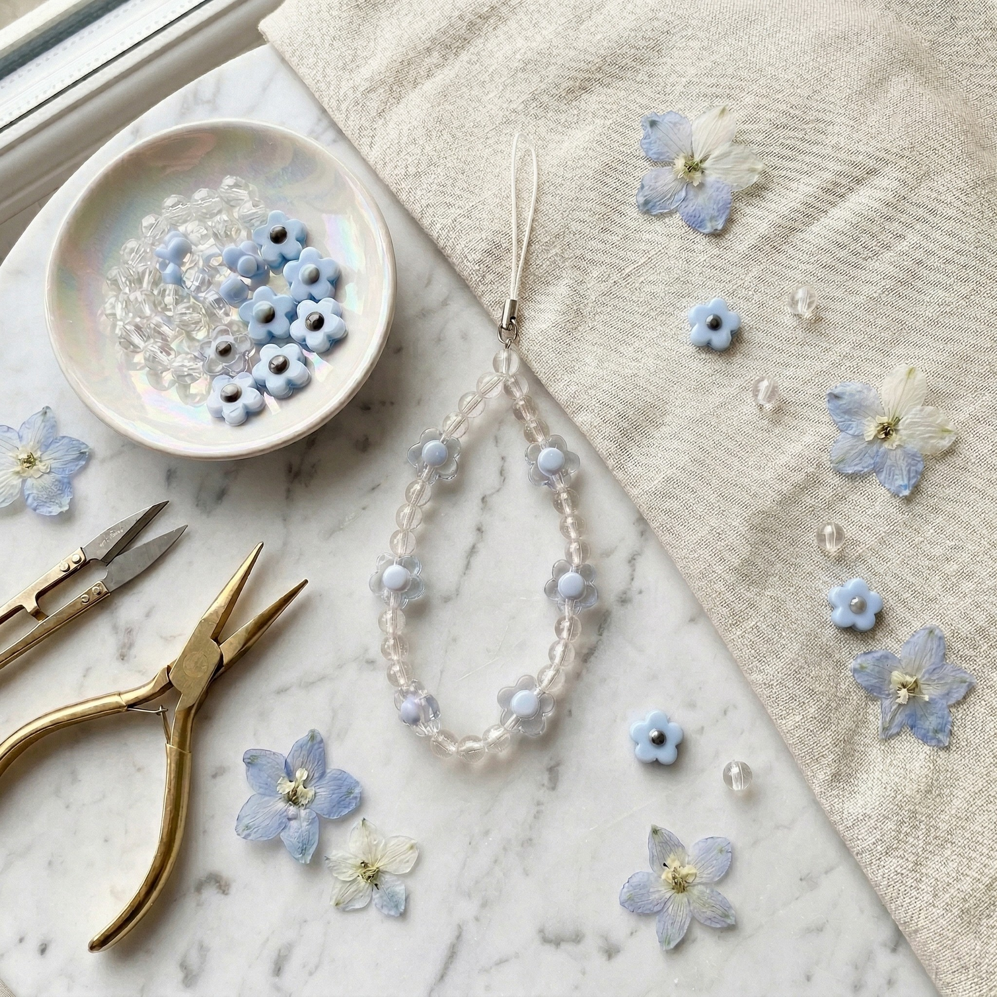 A delicate light blue and clear beaded phone charm shaped like small flowers, resting on a white marble surface next to loose beads, dried flowers, and golden crafting pliers.