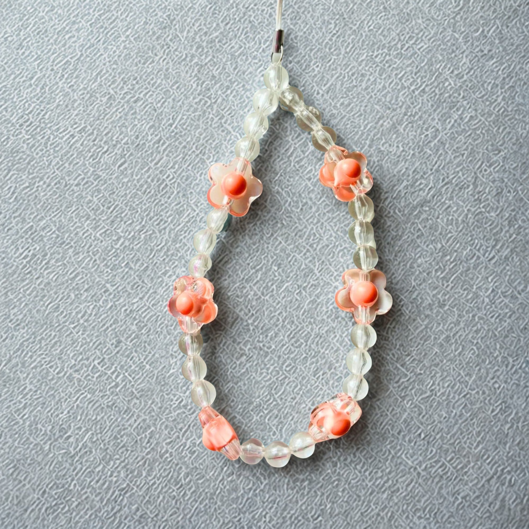 A beaded phone strap featuring peach-colored floral beads and clear round beads, resting on a textured grey surface.