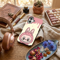 Glossy phone case featuring a cute pastel pink Labubu monster character in a bunny suit, laid on a desk next to rose gold headphones and blue flowers.