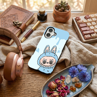 Glossy phone case featuring a cute sky blue Labubu monster character in a bunny suit, placed on a textured fabric alongside dried petals and a keyboard.