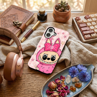Glossy phone case featuring a pink Labubu monster wearing a large bow on a pastel pink gingham background adorned with stars, laid on a desk next to rose gold headphones.