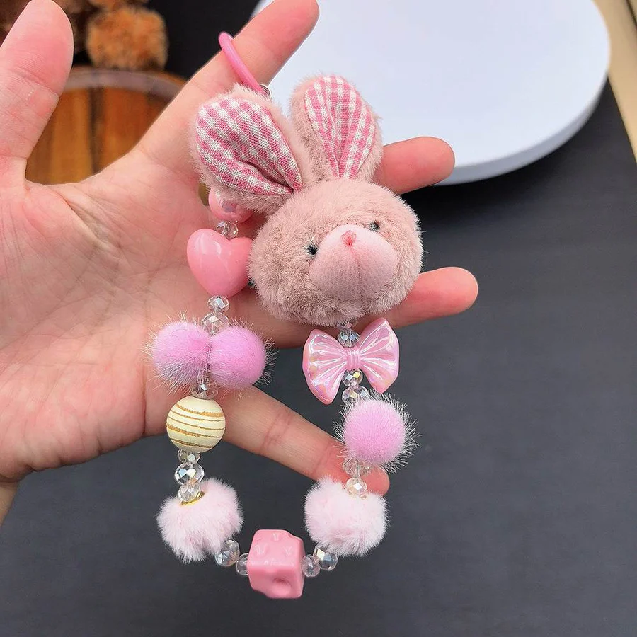 Hand holding a kawaii phone strap featuring a plush pink bunny head with gingham plaid ears, fluffy beads, a heart, and a pink dice charm.