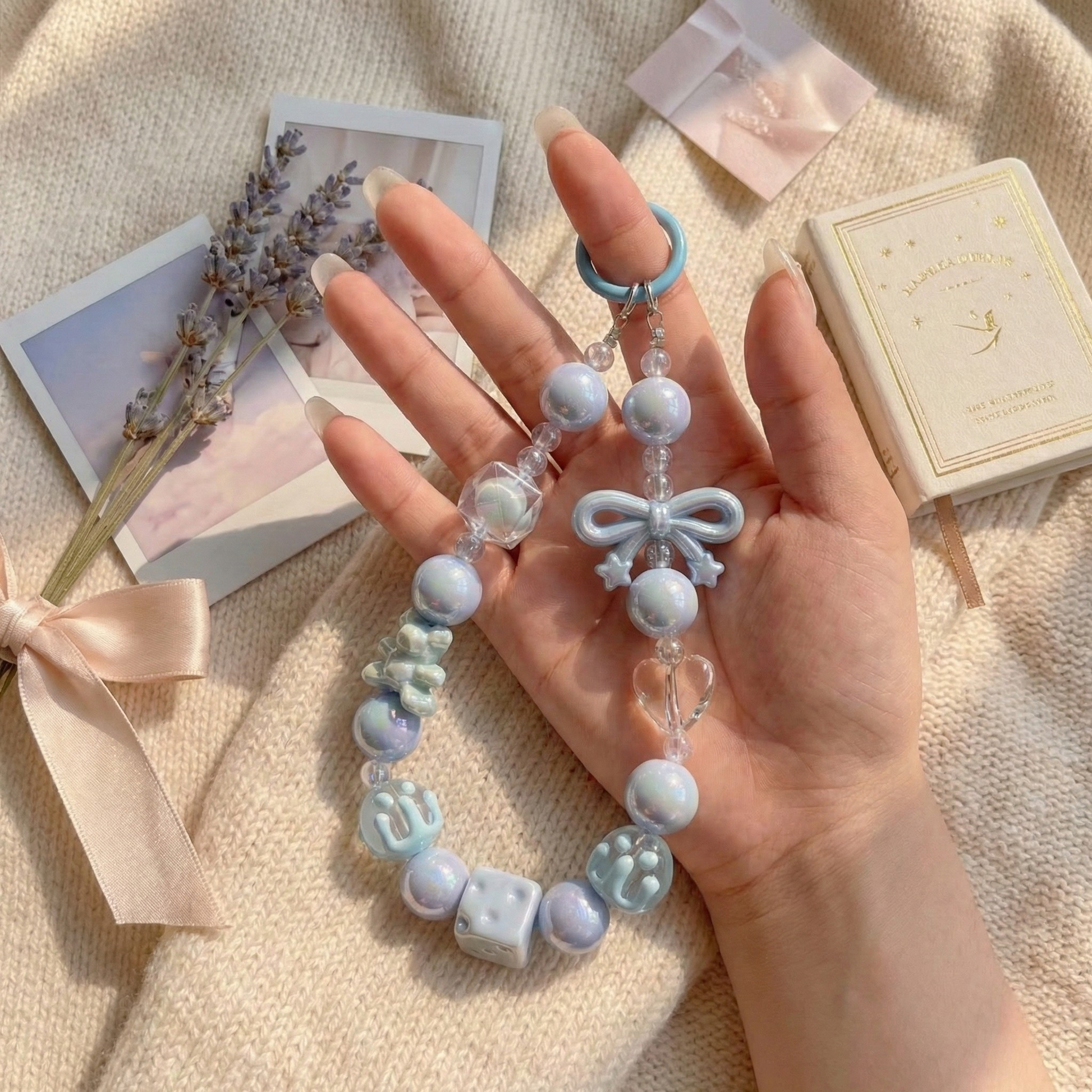 A hand holding a pastel blue beaded phone charm featuring iridescent pearls, a small bow, and clear heart accents against a soft, aesthetic background.