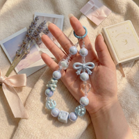 A hand holding a pastel blue beaded phone charm featuring iridescent pearls, a small bow, and clear heart accents against a soft, aesthetic background.