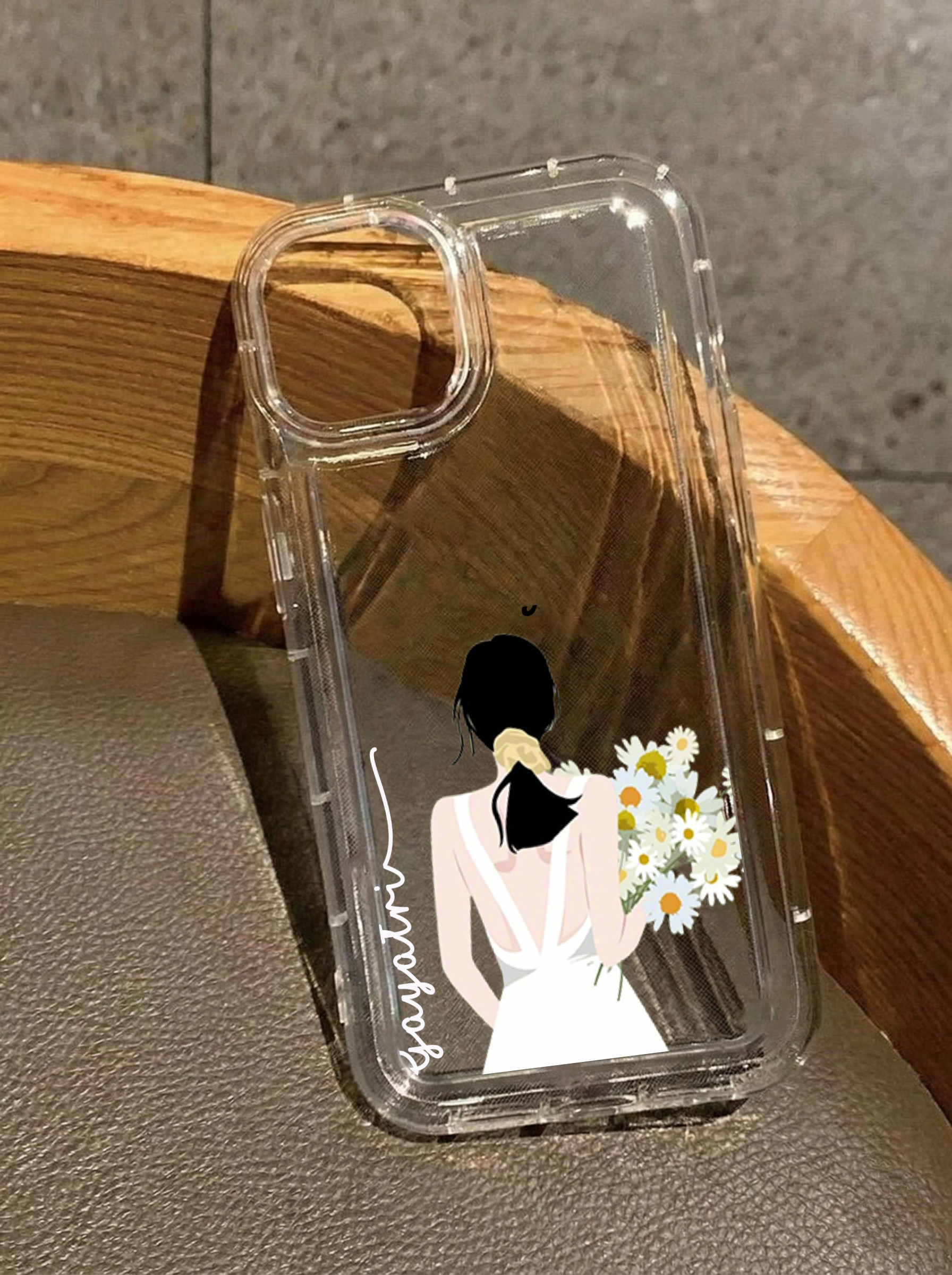 Clear iPhone 16 case featuring an illustration of a girl from behind in a white backless dress holding daisies, customized with the name Gayatri in vertical white text.