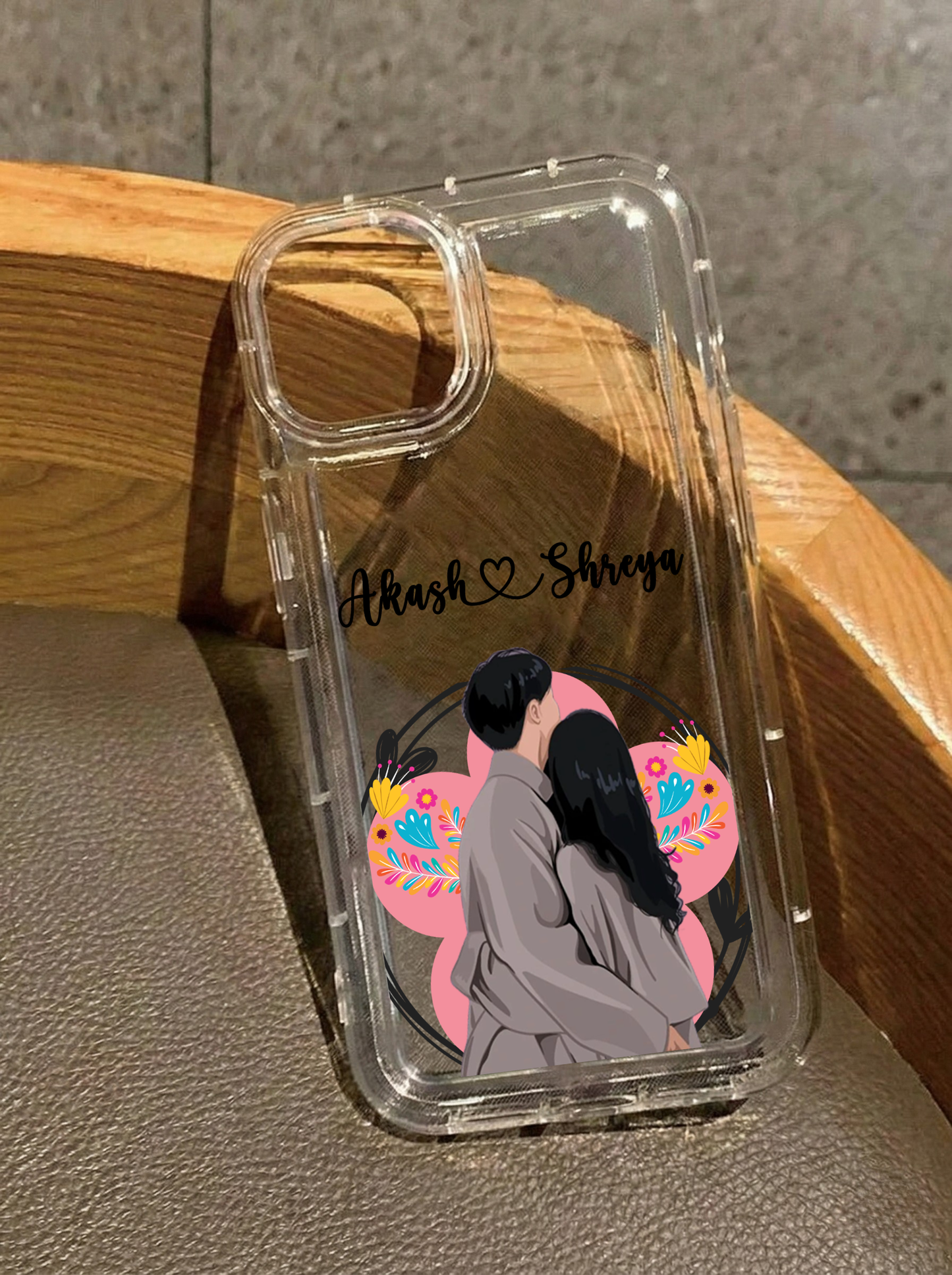 Clear iPhone 16 case featuring an illustration of a couple hugging viewed from behind against a pink floral circle, customized with the names Akash & Shreya in black script.
