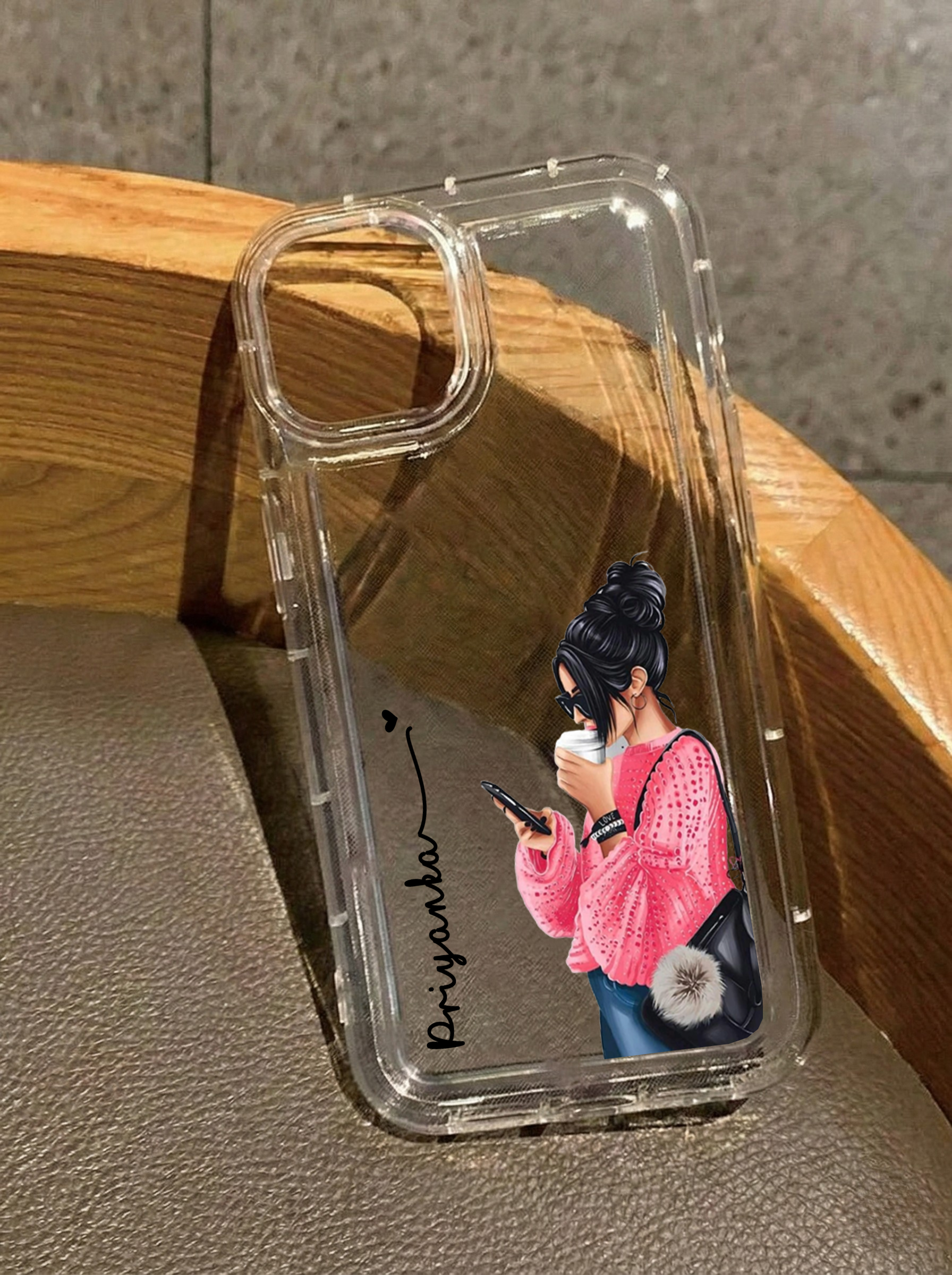 Clear iPhone 16 case showing an illustration of a girl with a messy bun and pink sweater holding a coffee cup and looking at her phone, customized with the name Priyanka in vertical black text.