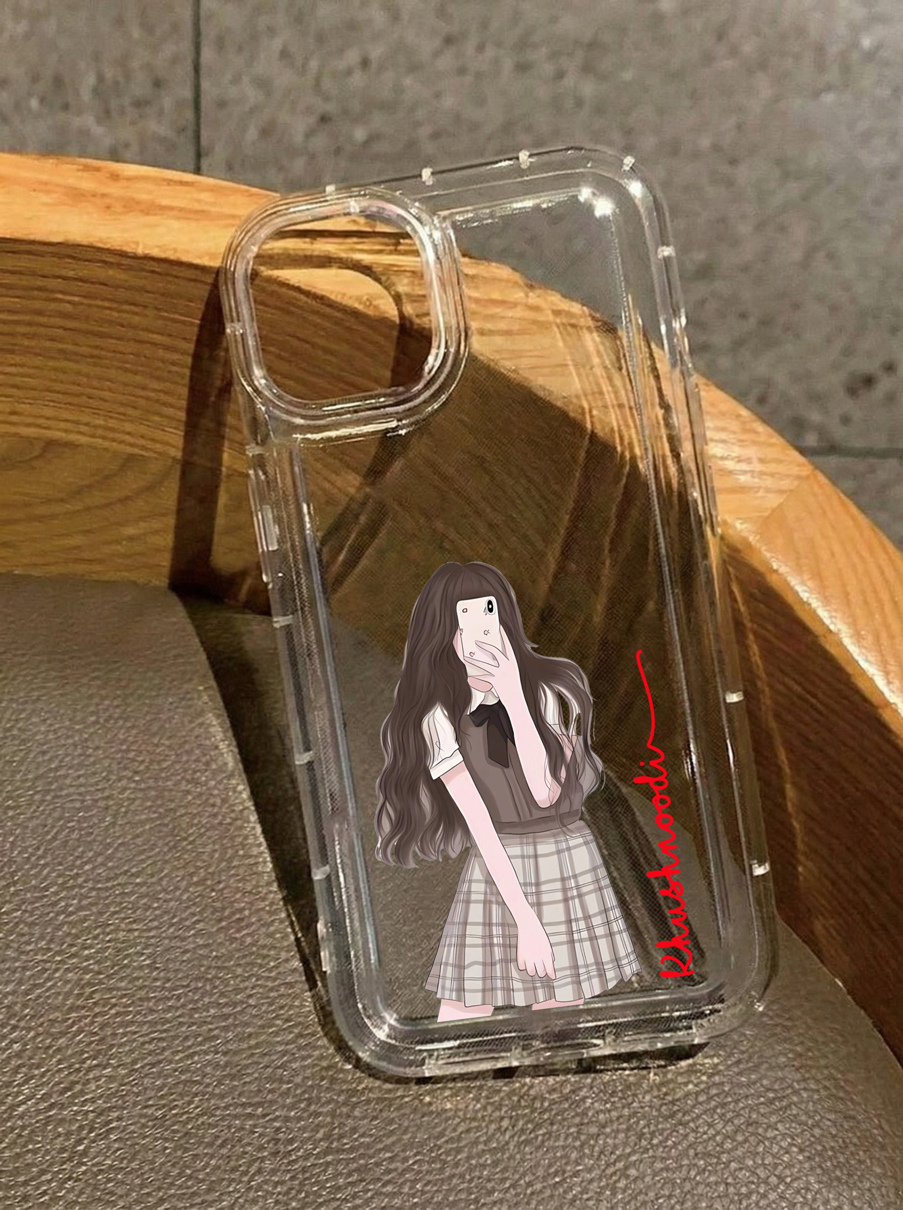 Clear iPhone 16 case featuring an illustration of a girl in a preppy plaid skirt taking a mirror selfie, customized with the name Khushnoodi in vertical red text.