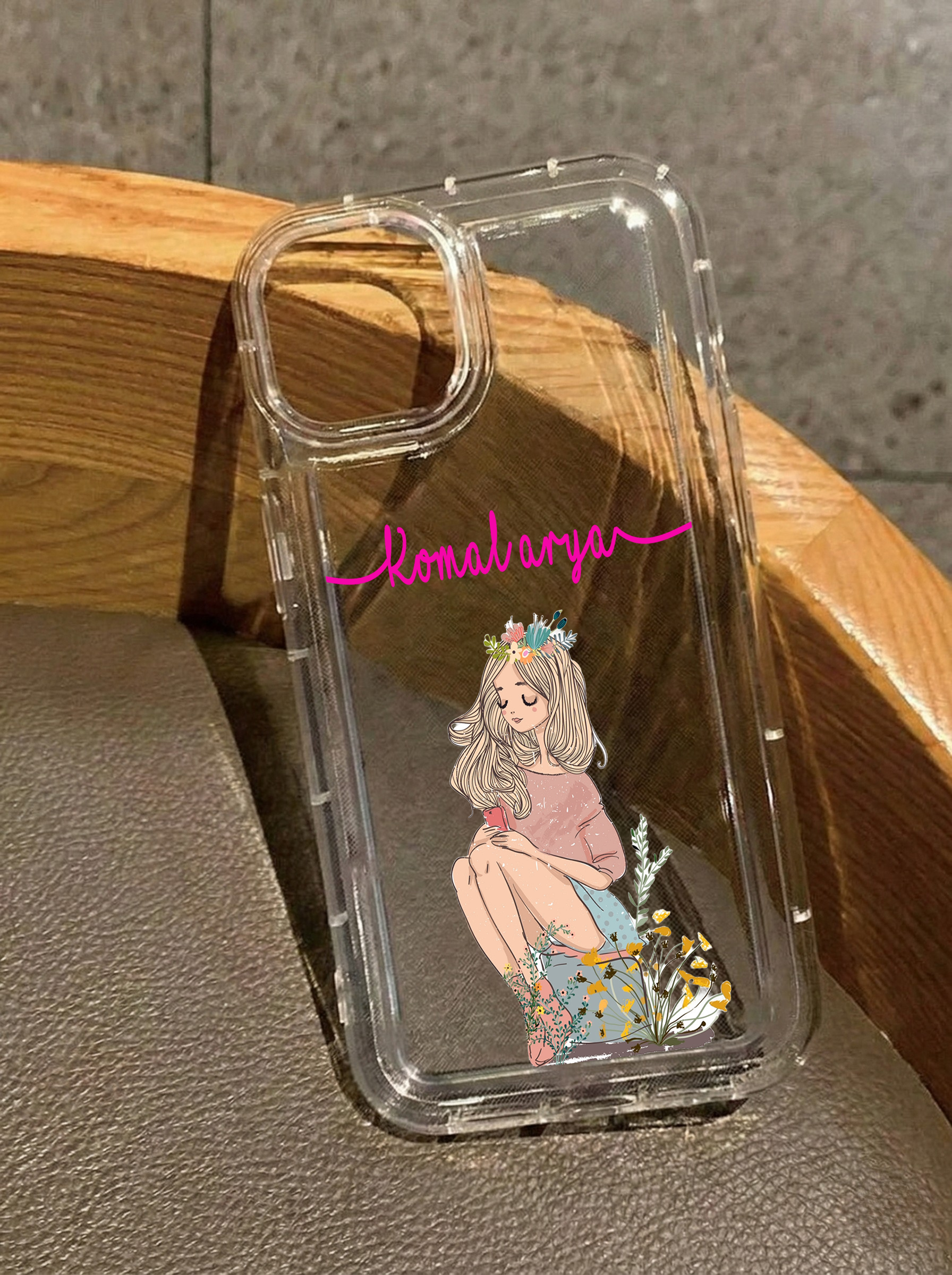 Clear iPhone 16 case featuring an illustration of a girl sitting down wearing a floral crown and holding a phone, customized with the name Komalarya in pink script.