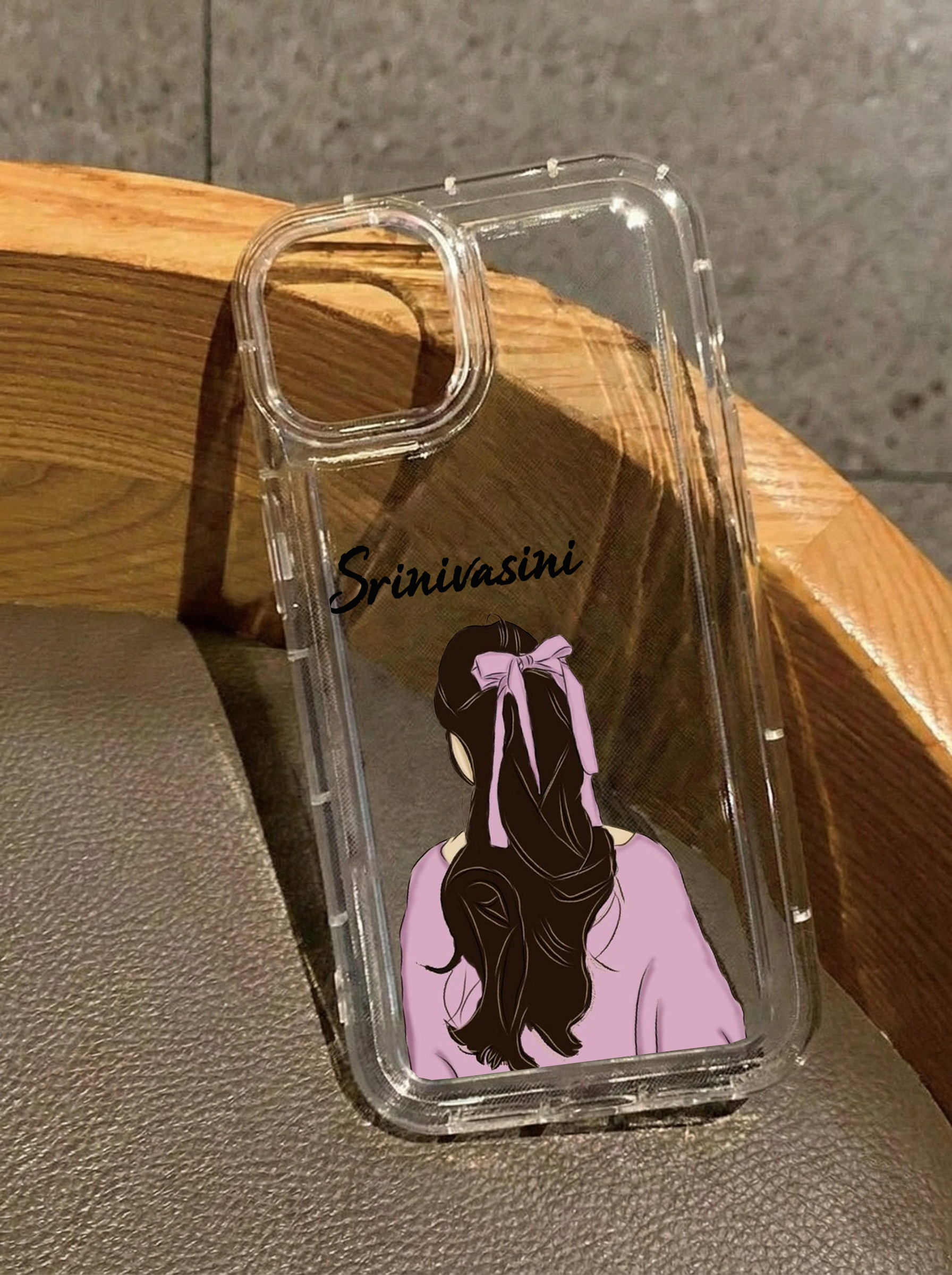 Clear iPhone 16 case showing an illustration from behind of a girl with long wavy hair wearing a pink bow and a pink top, customized with the name Srinivasini in black text.
