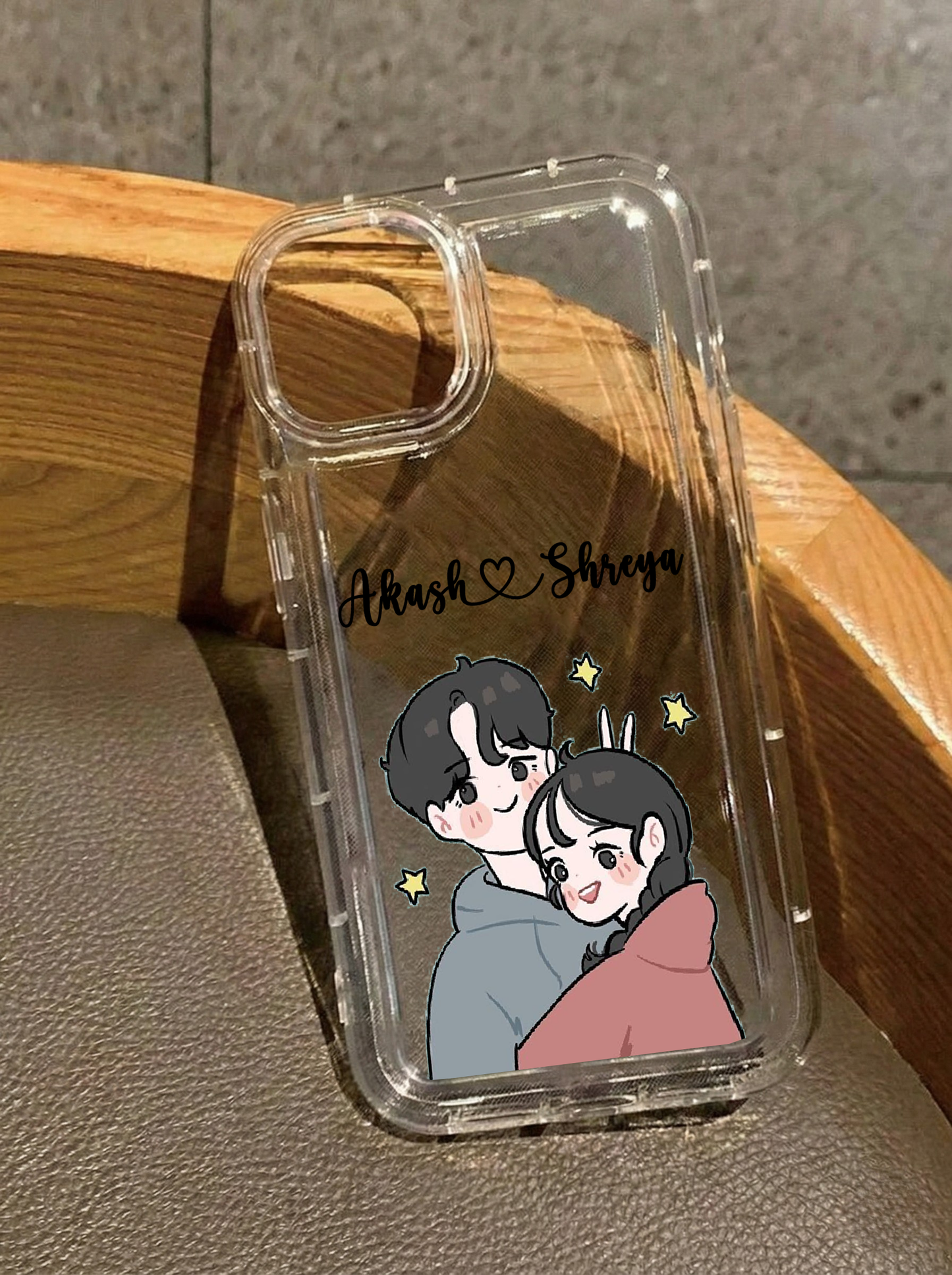 Clear iPhone 16 case displaying a cute anime-style illustration of a couple hugging surrounded by yellow stars, customized with the names Akash & Shreya in black script.