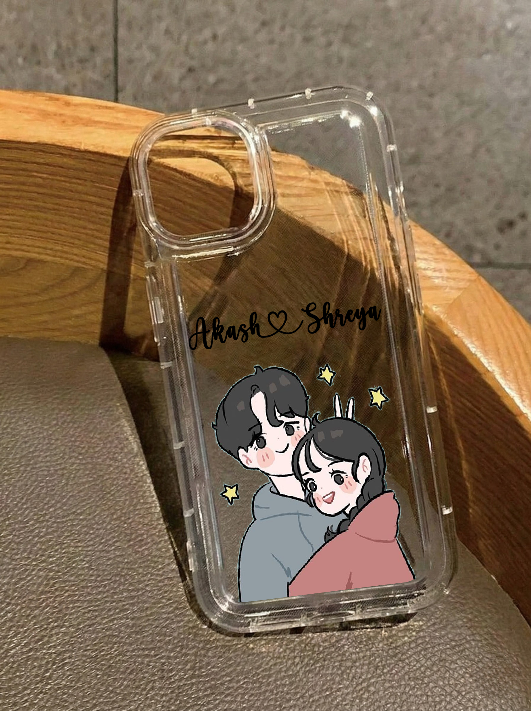 Clear iPhone 16 case displaying a cute anime-style illustration of a couple hugging surrounded by yellow stars, customized with the names Akash & Shreya in black script.