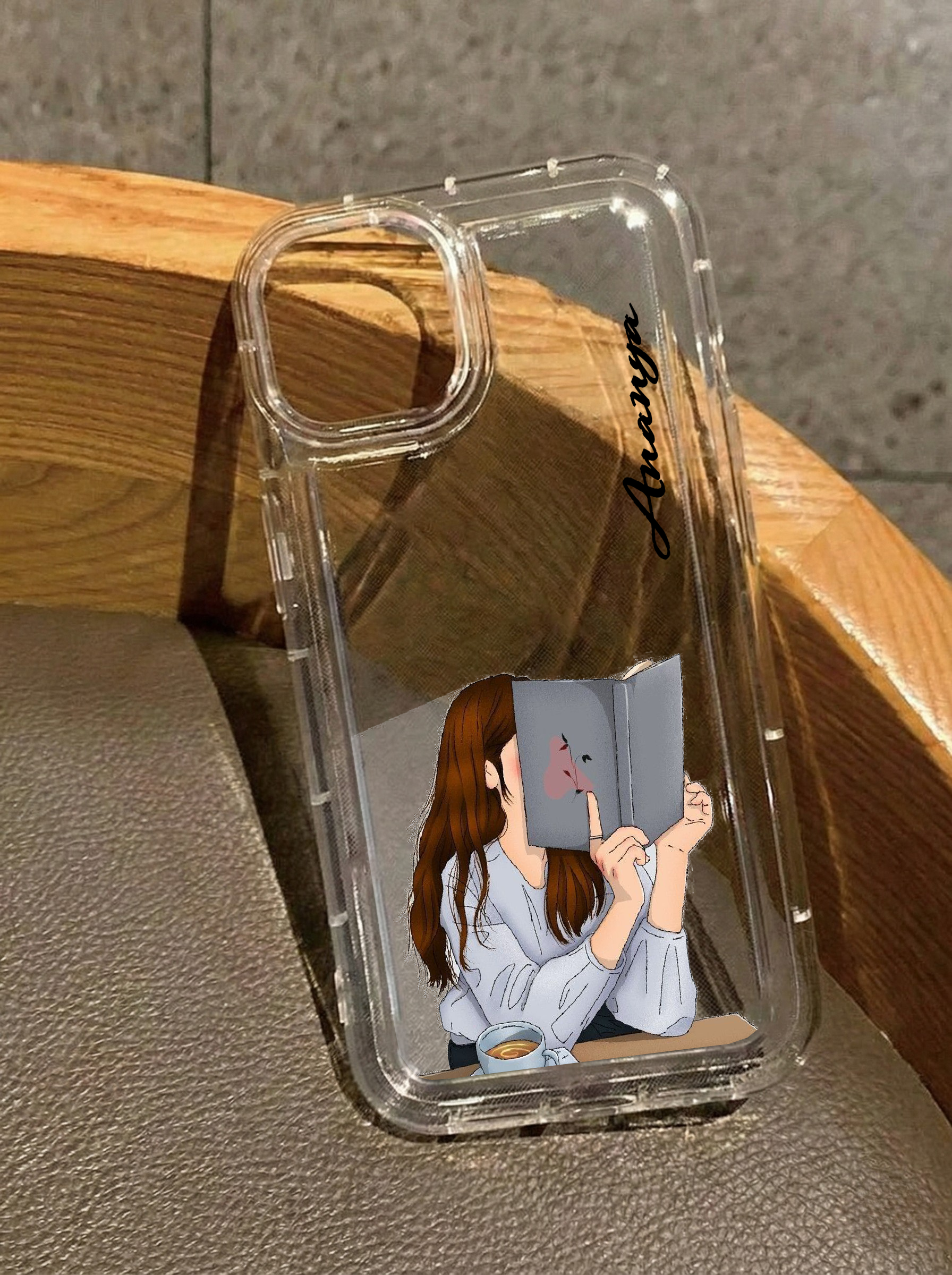 Clear iPhone 16 case featuring an illustration of a girl hiding her face behind a book with a coffee cup on the table, customized with the name Ananya in vertical black text.