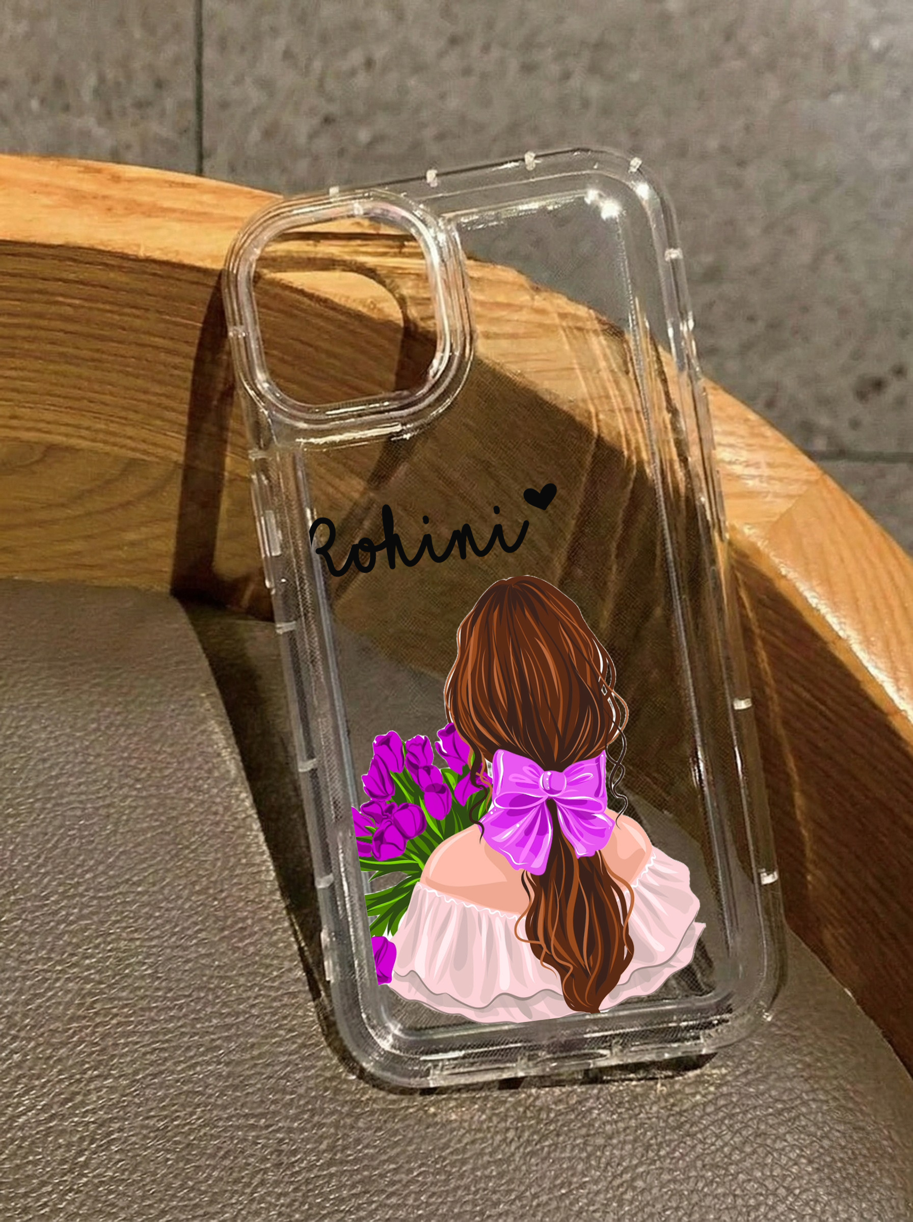 Clear iPhone 16 case featuring an illustration of a girl from behind with a pink hair bow holding purple tulips, customized with the name Rohini in black text.