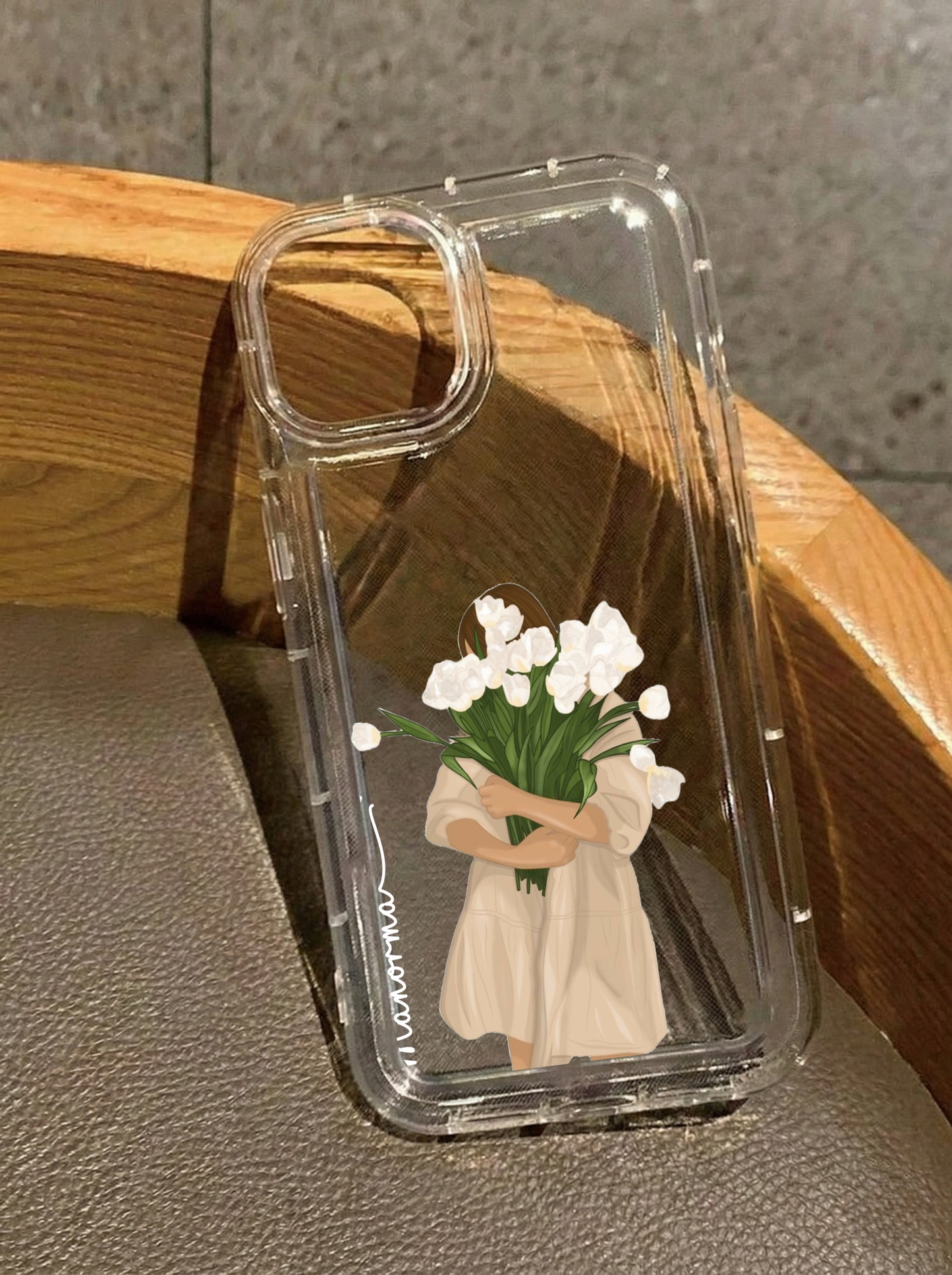 Clear iPhone 16 case displaying an illustration of a girl in a beige dress holding a large bouquet of white flowers, personalized with the name Manorma in vertical white text.