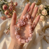 A hand displaying a soft pink beaded phone lanyard featuring iridescent pearl beads, a pink bow charm, and clear heart shapes with a romantic floral background.