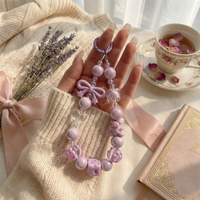 A hand holding a lavender purple beaded phone wristlet with iridescent pearls, a matching bow charm, and a teacup in the soft, aesthetic background.